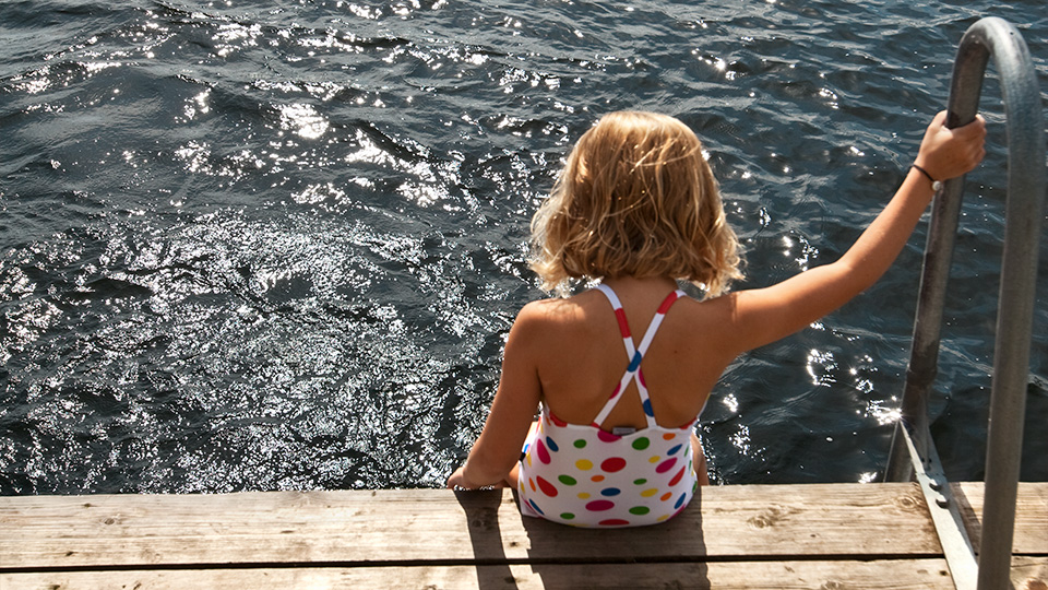 Ett barn i baddräkt sitter på en brygga och håller sig i räcket till stegen som går ner i vattnet.